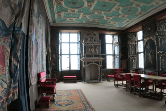 Ornate historic interior room at Bolsover Castle, featuring a decorative painted ceiling, large lead-light windows, wall tapestries, wooden furniture, and red upholstered chairs.