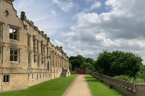 Exterior corridor view of Bolsover Castle in Derbyshire featuring sandstone walls, tall columned buttresses, lawn, and a pathway beside a stone balustrade with trees