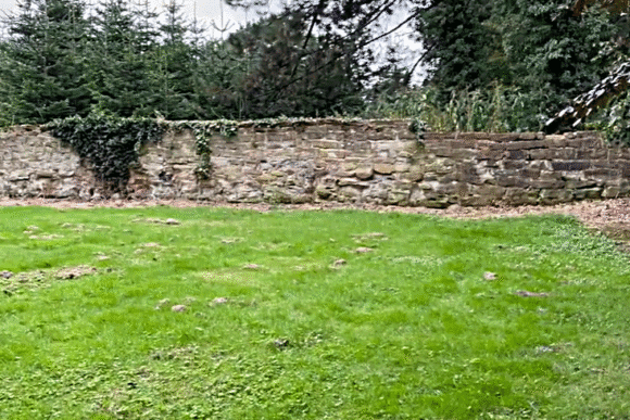 Stone wall and grassy archaeological area at the Aldborough Roman Site with scattered stones on the ground.