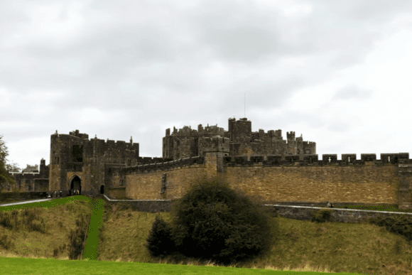 Wide view of Alnwick Castle with stone walls, towers, and surrounding greenery under an overcast sky.