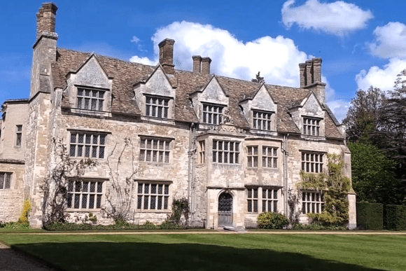 A large historic stone mansion with steep gabled roofs, tall chimneys, and mullioned windows, set beside manicured lawns at Anglesey Abbey.