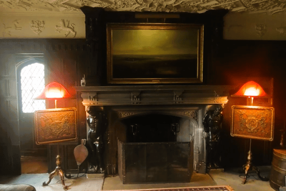 Historic interior room at Anglesey Abbey featuring a carved stone fireplace, dark wood paneling, a large framed painting, and two ornate lamps with red shades.