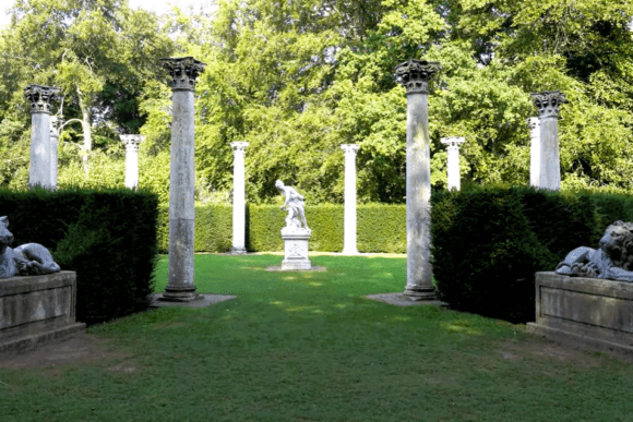 Garden at Anglesey Abbey featuring stone columns, lion sculptures, and a classical statue surrounded by greenery.