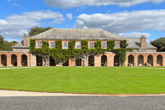 Historic courtyard and brick colonnade at Antony House in Cornwall, facing a central lawn under a bright sky.