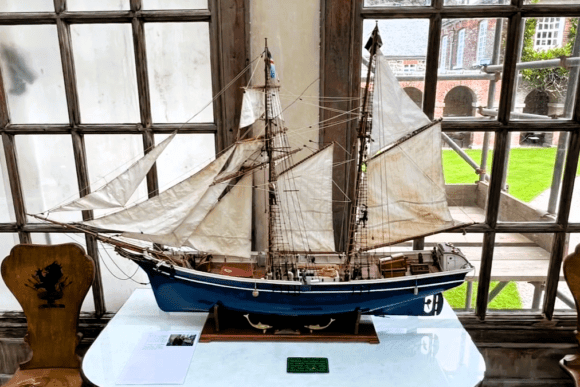 Detailed model sailing ship displayed at Antony House in Cornwall, set on a table in front of large historic windows.
