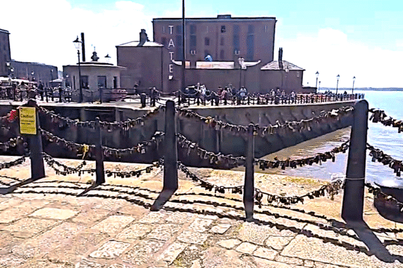 Waterfront area at Royal Albert Dock Liverpool with historic brick buildings, railings covered in padlocks, and visitors walking beside the water.