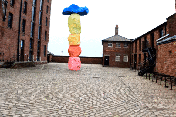 Courtyard at Royal Albert Dock Liverpool with historic red-brick warehouses and a tall multicoloured stone sculpture.