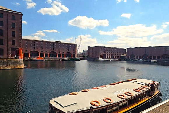 Waterfront view of Royal Albert Dock Liverpool with historic brick warehouses, boats on the water, and a large wheel in the background.