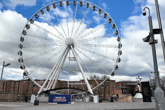 Large Ferris wheel known as the Wheel of Liverpool standing beside waterfront buildings under a partly cloudy sky.