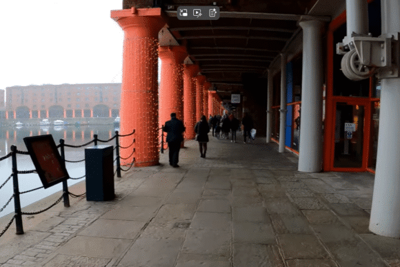 Covered walkway at Royal Albert Dock Liverpool with red columns, waterfront railings, and people walking beside the dock.