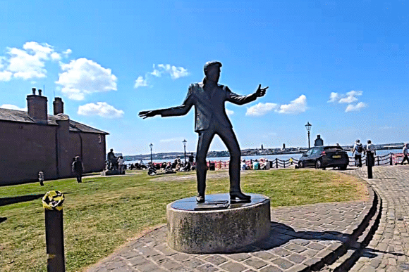 Bronze statue of a performing figure with arms outstretched on a plinth at Liverpool’s waterfront, with people and the River Mersey in the background.