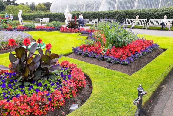 Colourful flower beds and manicured lawns at Kew Gardens in London, with vibrant seasonal blooms, garden sculptures, benches, and the iconic Palm House glasshouse in the background.