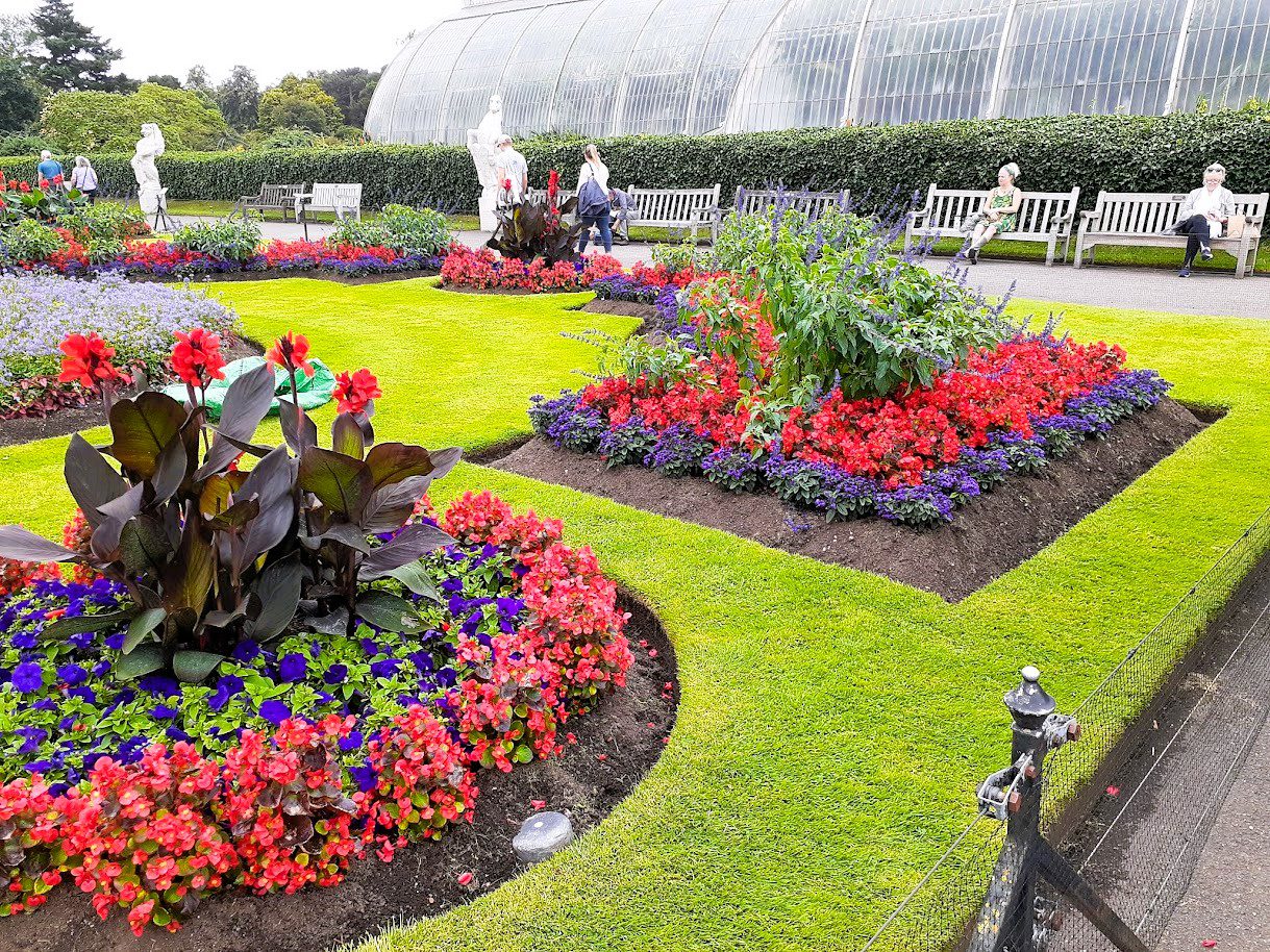 Colourful flower beds and manicured lawns at Kew Gardens in London, with vibrant seasonal blooms, garden sculptures, benches, and the iconic Palm House glasshouse in the background.