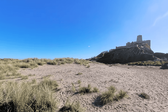 Bamburgh Beach with golden sand dunes and the iconic Bamburgh Castle perched on a rocky outcrop overlooking the Northumberland coastline under a clear blue sky.