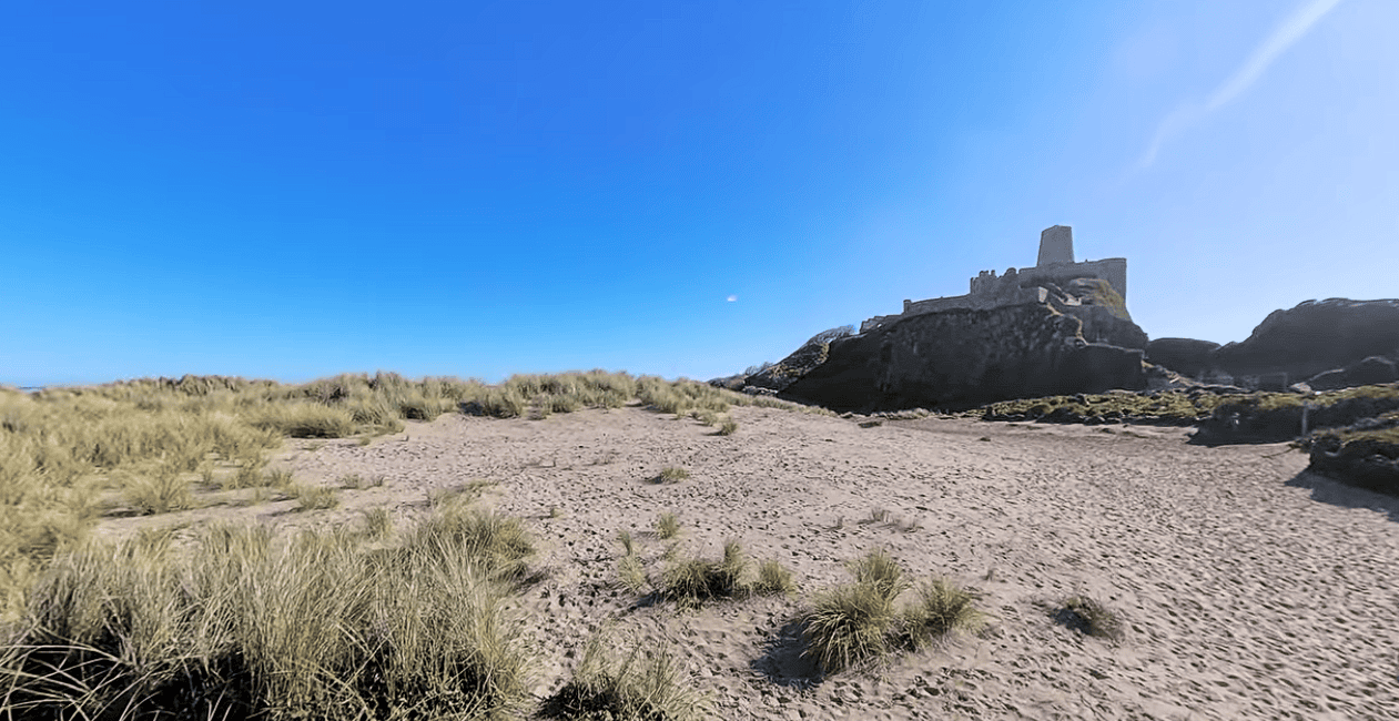 Bamburgh Beach with golden sand dunes and the iconic Bamburgh Castle perched on a rocky outcrop overlooking the Northumberland coastline under a clear blue sky.