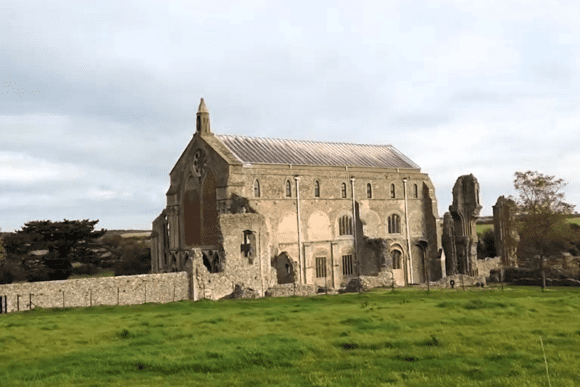 Ruins of a medieval stone priory church under a cloudy sky, standing beside a grassy field with surrounding boundary walls and trees.