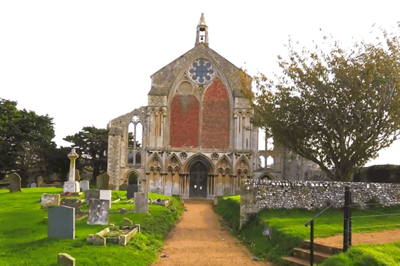 Partially ruined medieval priory church with a bell-cote steeple, stone arch windows, and red brick infill, surrounded by gravestones and a grassy churchyard under a cloudy sky.