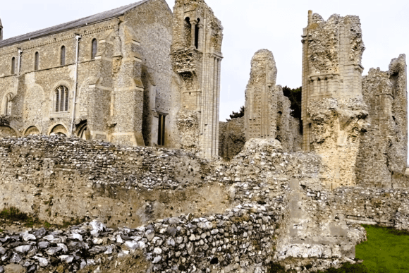 Stone ruins of a medieval priory showing tall arched window openings, textured flint-and-stone walls, and a small bell-cote on the roofline, with grassy ground in the foreground.