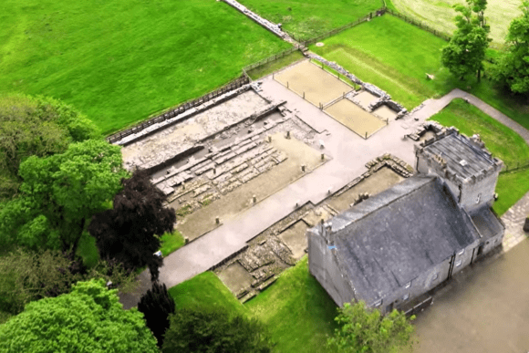 Aerial view of ancient Roman fort ruins showing rectangular stone foundations, exposed internal building layouts, fenced excavation areas, and surrounding green fields with trees.