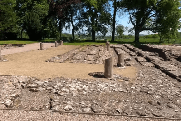 Archaeological ruins of Birdoswald Roman Fort showing exposed stone foundations, timber post remnants marking building outlines, gravel visitor paths, and surrounding woodland and green landscape.