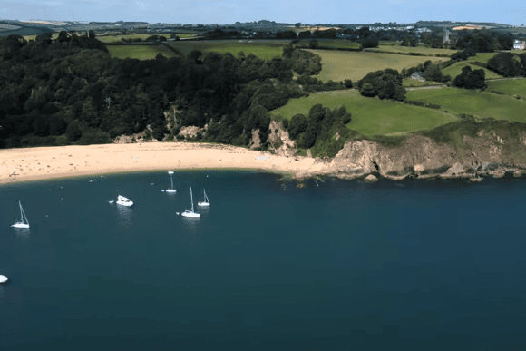Aerial view of Blackpool Sands in Devon with golden beach, turquoise bay, anchored sailboats, and lush green countryside surrounding the picturesque South Devon coastline.