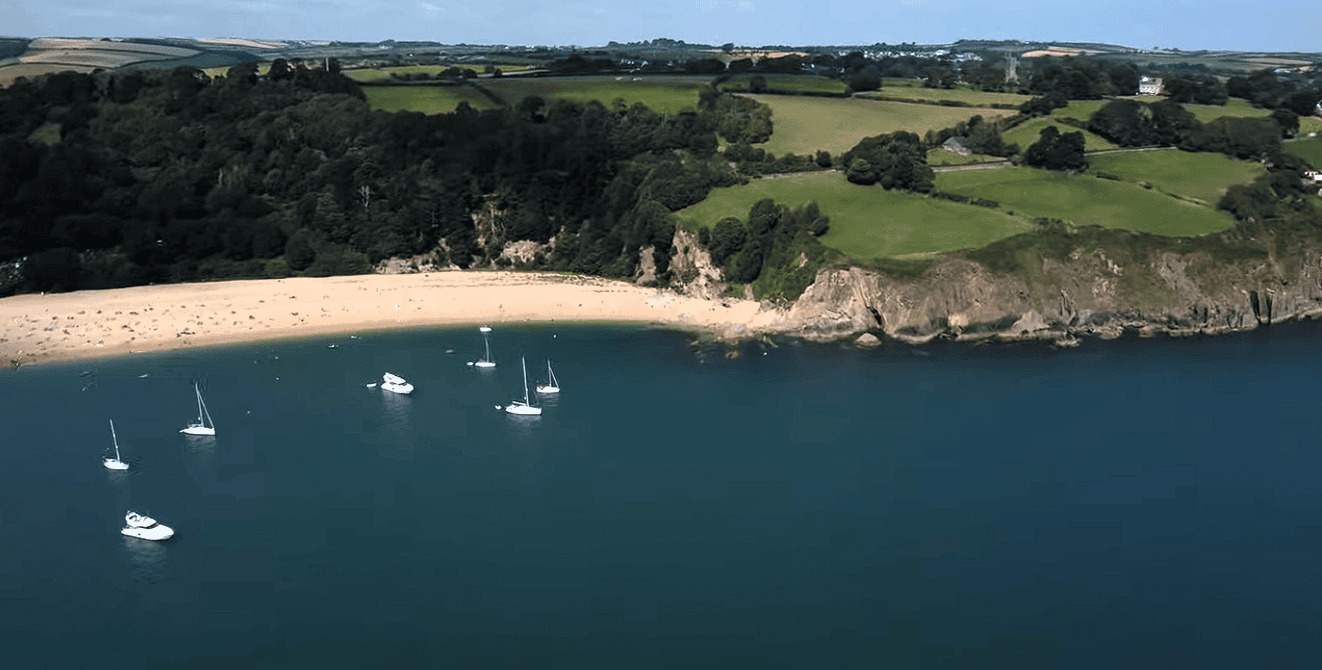 Aerial view of Blackpool Sands in Devon with golden beach, turquoise bay, anchored sailboats, and lush green countryside surrounding the picturesque South Devon coastline.