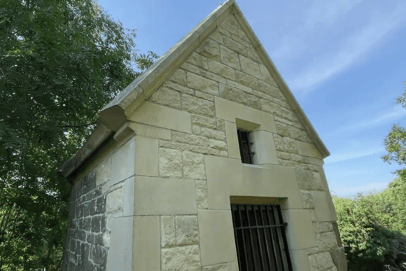 Small stone-built Cundy House at Bolsover, showing a gabled roof and two narrow window openings with iron bars, set in a wooded landscape under a blue sky.