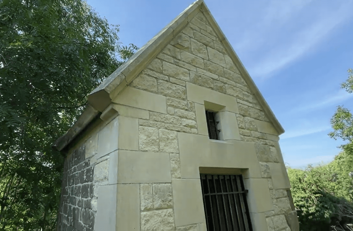 Small stone-built Cundy House at Bolsover, showing a gabled roof and two narrow window openings with iron bars, set in a wooded landscape under a blue sky.
