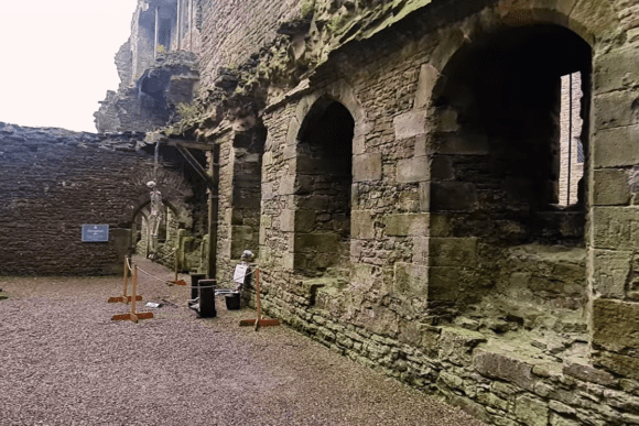 Bolton Castle interior ruins featuring weathered stone walls with large arched openings and windows, a gravel courtyard floor, and a roped walkway guiding visitors toward a carved medieval doorway.