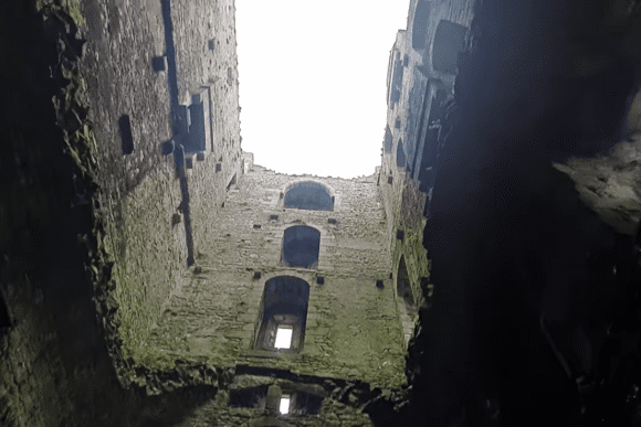 Interior view of Bolton Castle, showing tall moss-covered stone walls and a vertical line of arched window openings reaching toward a bright open sky above the ruined courtyard.