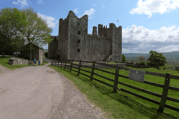 Bolton Castle, a large medieval stone fortress with corner towers, narrow windows, and a flag on the roof, beside a rural path and wooden fence, set against green hills and a partly cloudy sky.
