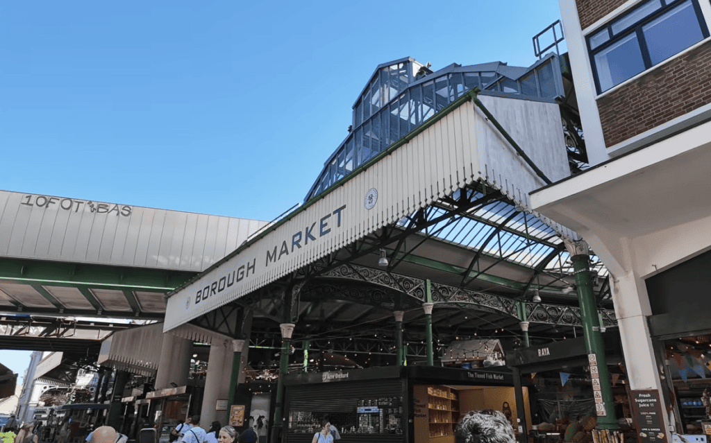Main entrance to Borough Market in London featuring its iconic Victorian ironwork, glass roof, and bustling food market beneath a clear blue sky.