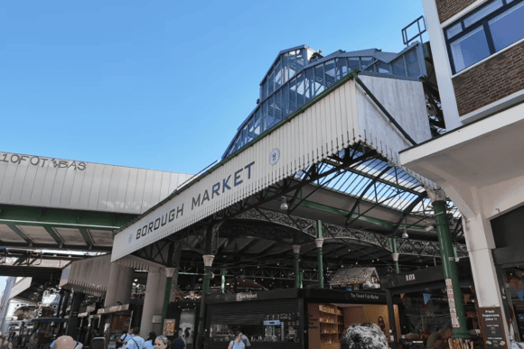 Main entrance to Borough Market in London featuring its iconic Victorian ironwork, glass roof, and bustling food market beneath a clear blue sky.