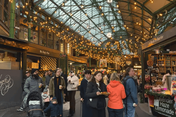 Crowds of visitors exploring Borough Market in London beneath a Victorian glass roof with illuminated string lights, artisan food stalls, and gourmet vendors.
