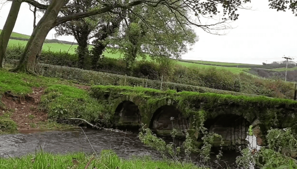 Historic Bow Bridge in Bolton Abbey, a moss-covered triple-arched stone footbridge crossing the River Wharfe, surrounded by woodland and green countryside in North Yorkshire, England.