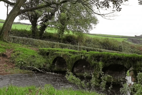Historic Bow Bridge in Bolton Abbey, a moss-covered triple-arched stone footbridge crossing the River Wharfe, surrounded by woodland and green countryside in North Yorkshire, England.