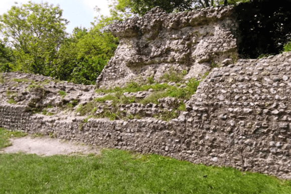 Low stone defensive walls of Bramber Castle ruins built from rounded flint and rubble, partially covered with grass and moss, with trees and green foliage behind, and a grassy foreground leading to the base of the medieval wall.