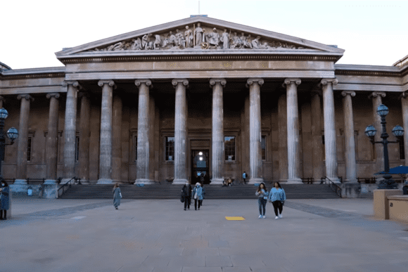 The front façade of the British Museum featuring a large triangular pediment with sculpted figures, a long row of tall classical columns, wide stone steps leading to the entrance, and visitors walking across the open forecourt under a pale daylight sky.