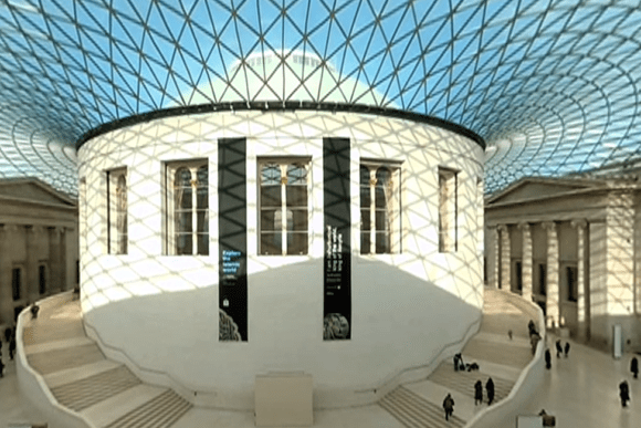 The Great Court of the British Museum with a vast geometric glass roof forming a lattice pattern above a large circular reading room at the centre. Sunlight casts patterned shadows on the floor, while visitors walk along curved staircases and across the open court inside the historic museum hall.
