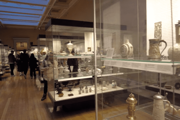 Visitors walking through a British Museum gallery with glass display cabinets on both sides containing silver and metal artefacts, including ornate jugs, plates, urns, and vessels. The museum hall features a wooden floor and a skylit ceiling above.