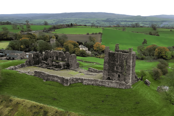 Elevated view of Brough Castle ruins showing a tall rectangular stone keep, surrounding curtain walls, and the remains of inner buildings set on a grassy rise, with patchwork green fields, trees, and rolling hills of the Eden Valley stretching into the distance.