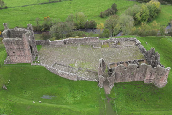 Aerial view of the ruins of Brough Castle showing rectangular stone walls, a tall surviving tower, collapsed sections of masonry, and a large open courtyard, set within green pastureland beside a winding river and trees.