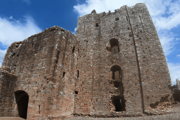Tall stone towers and curtain walls of Brougham Castle rising from a gravel courtyard, showing arched doorways, narrow window openings, weathered masonry, and patches of vegetation on the walls beneath a blue sky with scattered clouds.
