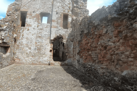 Interior courtyard view at Brougham Castle showing weathered red and grey stone walls, narrow rectangular window openings, and a small arched doorway leading deeper into the ruins under a partly cloudy sky.