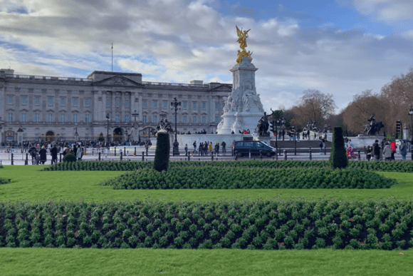 Wide view of Buckingham Palace with the Victoria Memorial in the foreground, showing manicured gardens, crowds of visitors, and the palace façade under a partly cloudy sky.