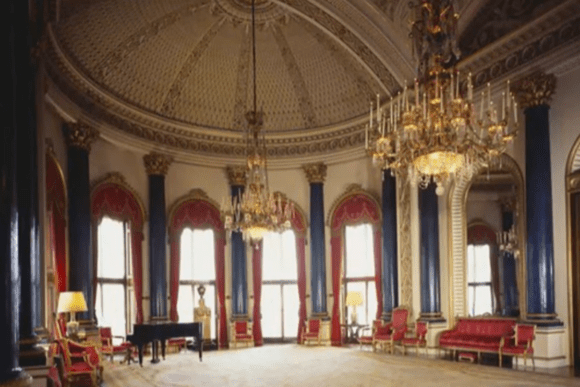 Ornate interior room at Buckingham Palace featuring a domed ceiling, grand crystal chandeliers, tall blue-and-gold columns, red upholstered seating, gilded mirrors, and tall windows draped with red curtains.