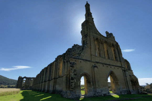 Ruined stone façade and arches of Byland Abbey standing in open countryside, backlit by the sun against a clear blue sky.