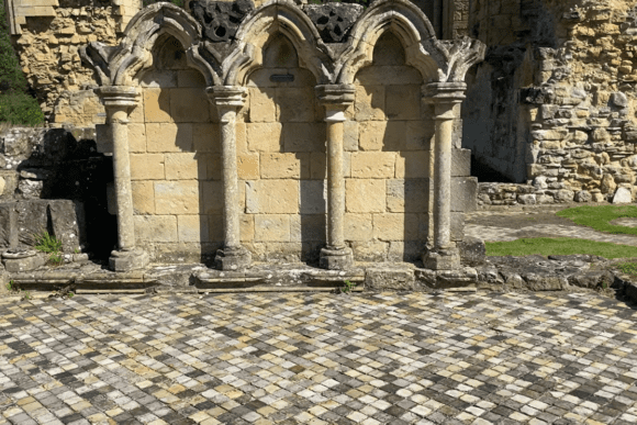 Row of carved stone columns and pointed arches at Byland Abbey, set against weathered masonry and a patterned stone courtyard.