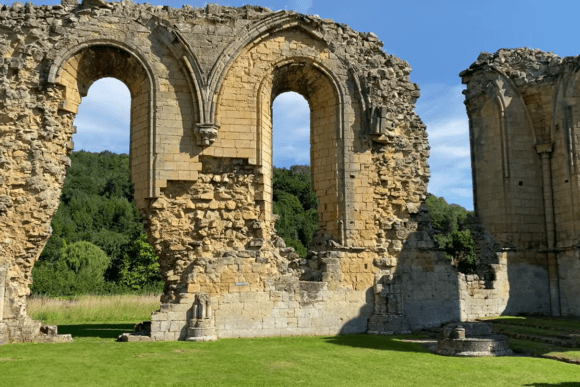Tall stone arches and window openings of Byland Abbey ruins standing on a grassy lawn, with wooded hills visible through the openings.