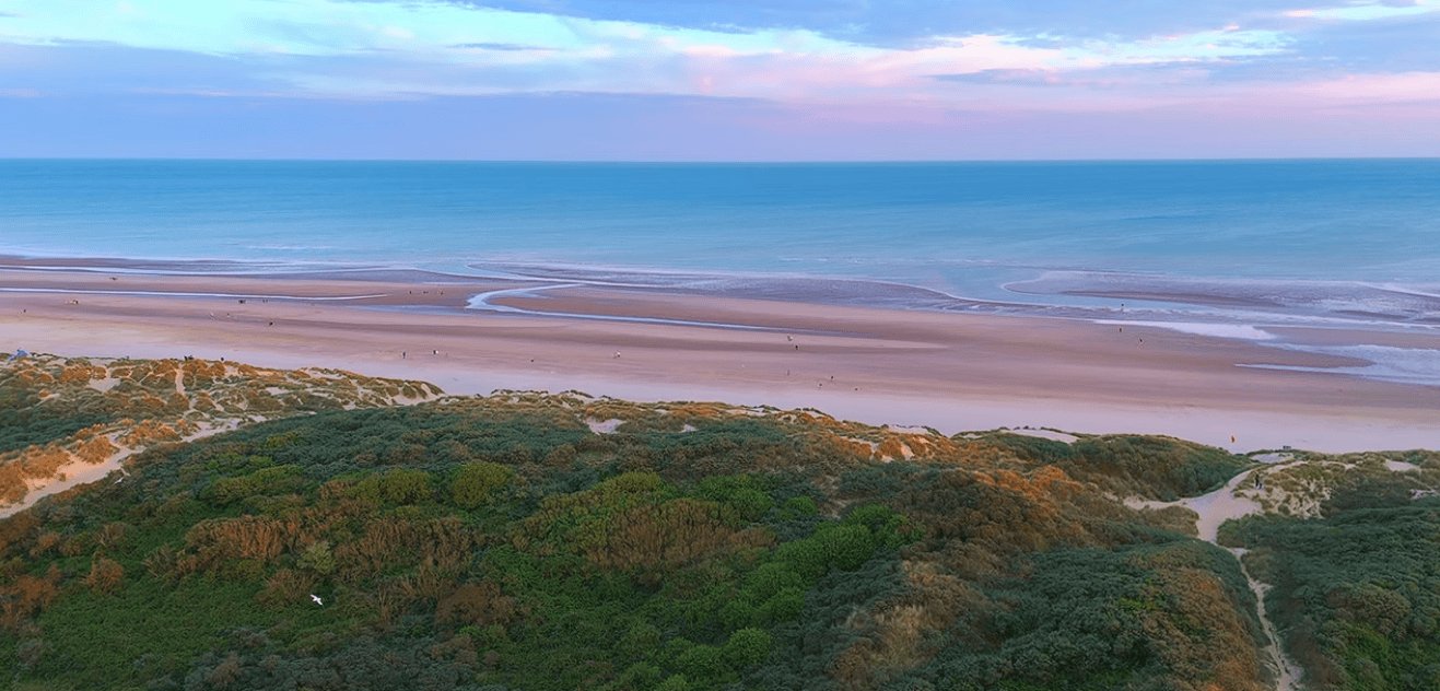 Aerial view of Camber Sands featuring sweeping golden sand dunes, wide sandy beach, shallow tidal channels, and the English Channel under a colorful evening sky in East Sussex.